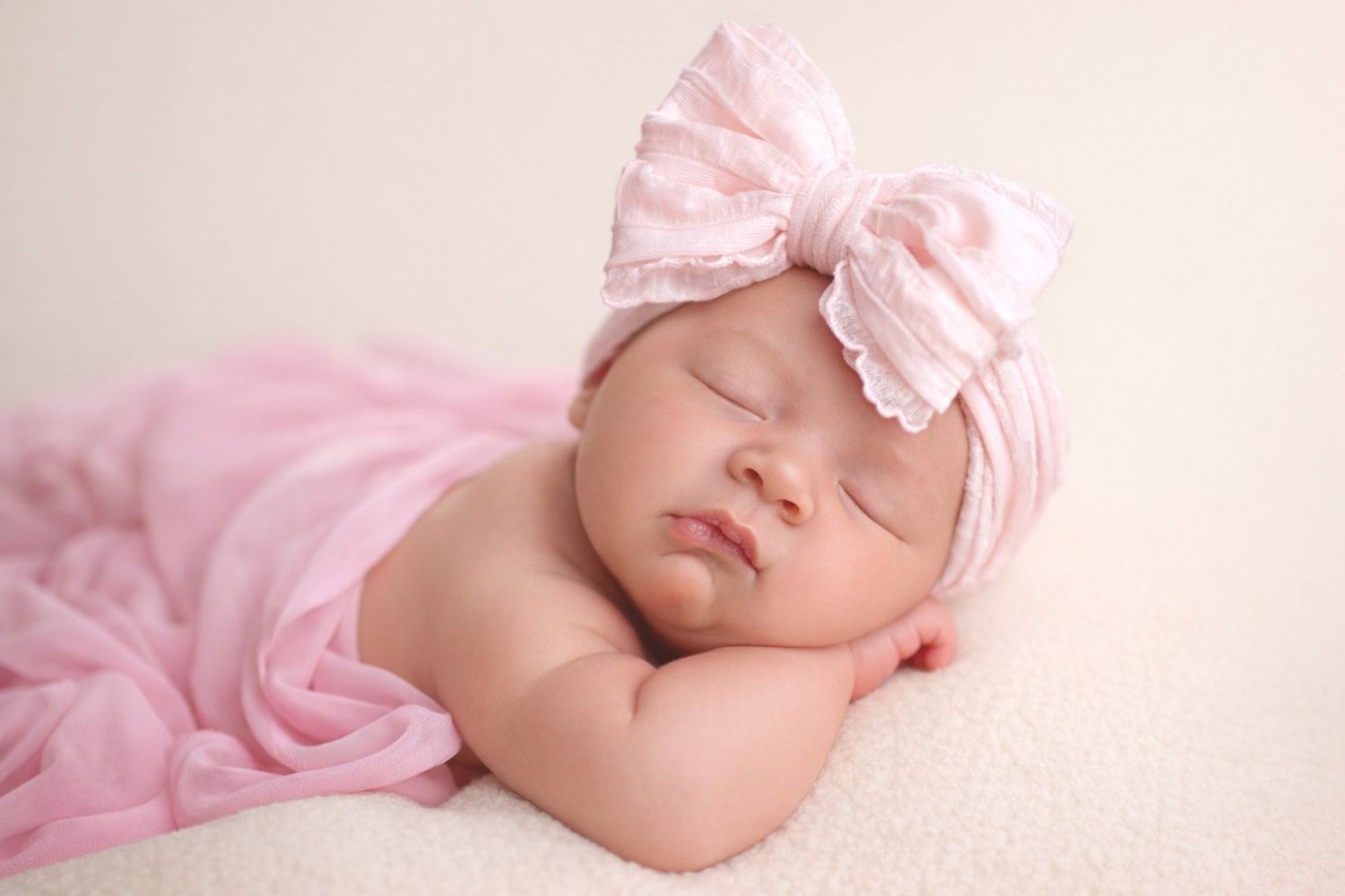 A sleeping newborn baby wrapped in soft pink fabric with a matching headband bow.