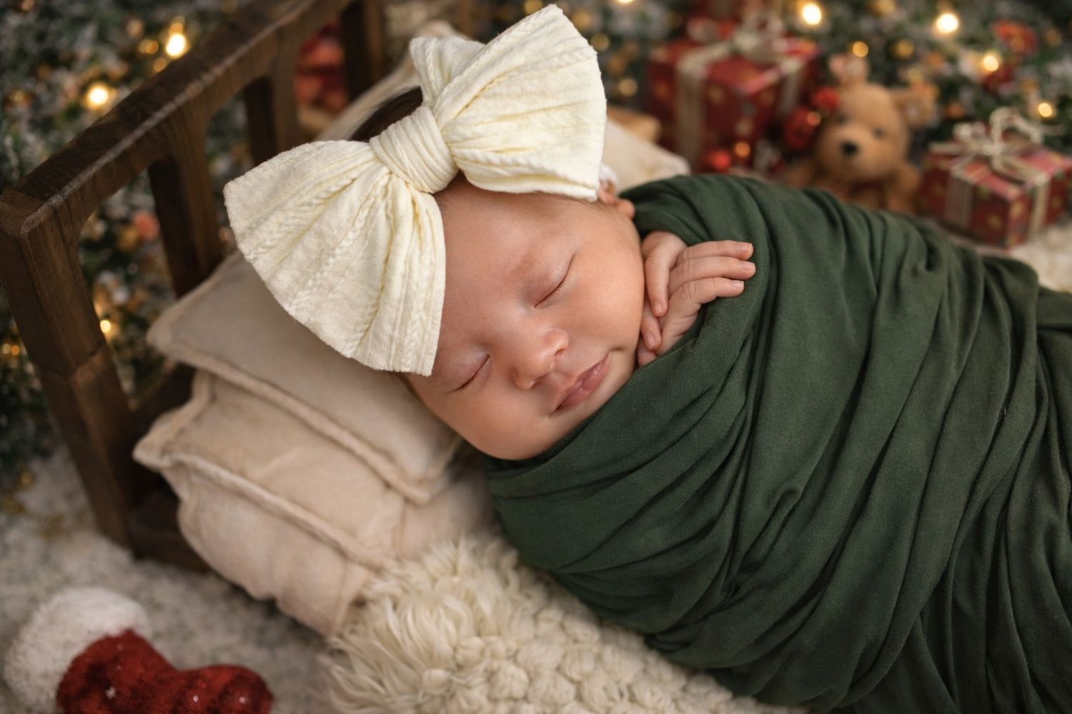 Baby in cream bow and green swaddle lying near Christmas decorations and twinkling lights.