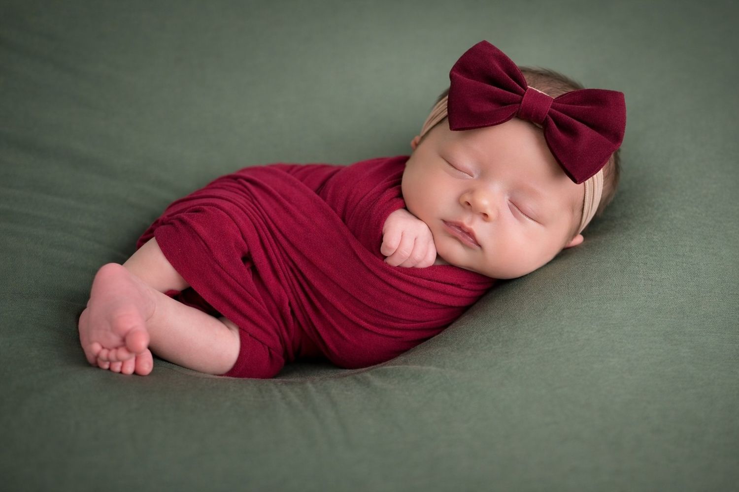 A sleeping baby wearing a burgundy onesie lies on a soft green blanket in a peaceful pose.