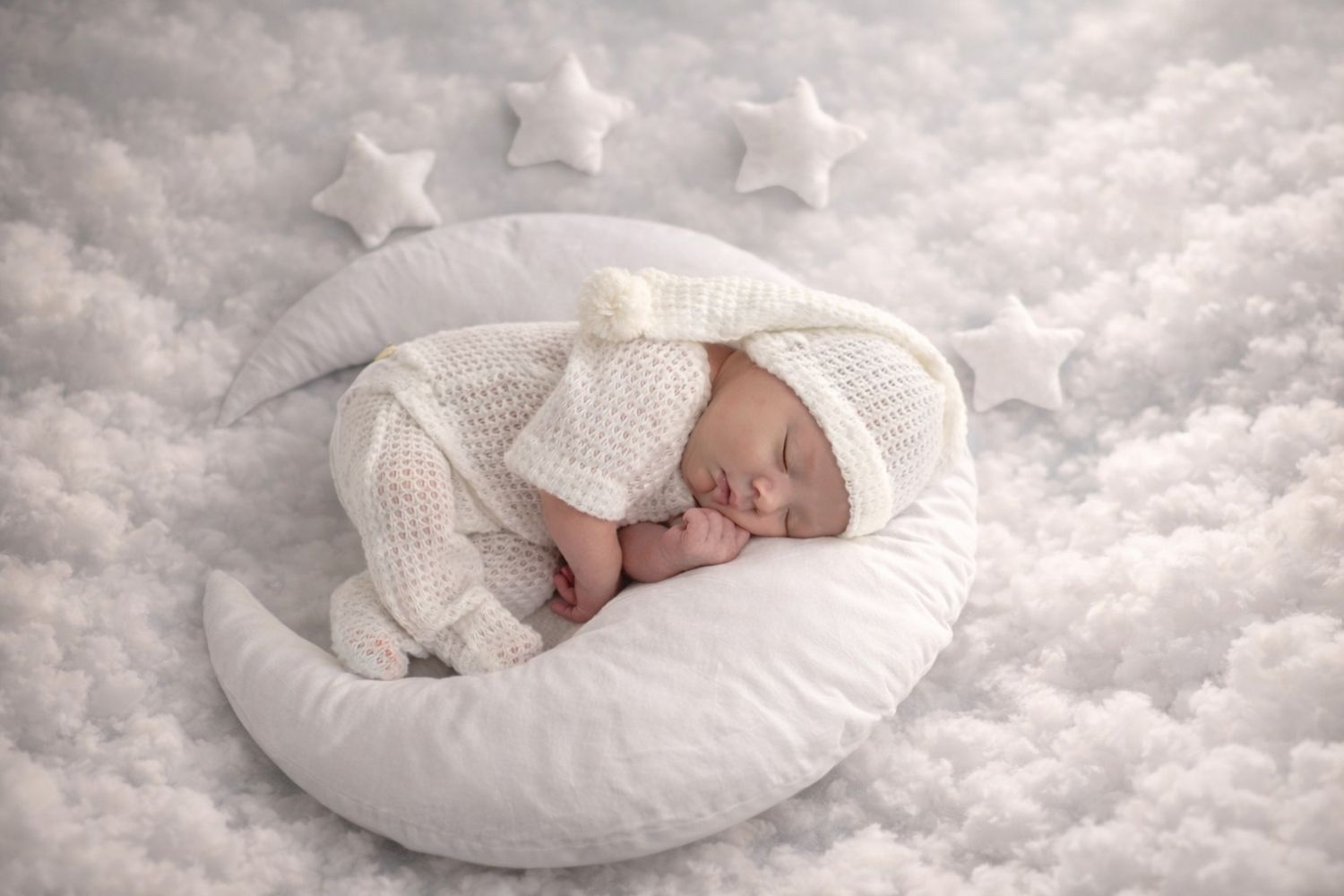 A newborn baby sleeping peacefully on a white cushioned surface with soft textured fabric in the background.