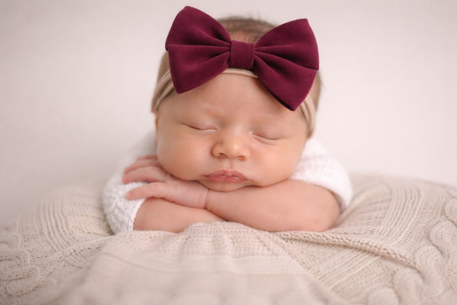 A sleeping baby wearing a burgundy bow headband rests peacefully on white textured fabric.