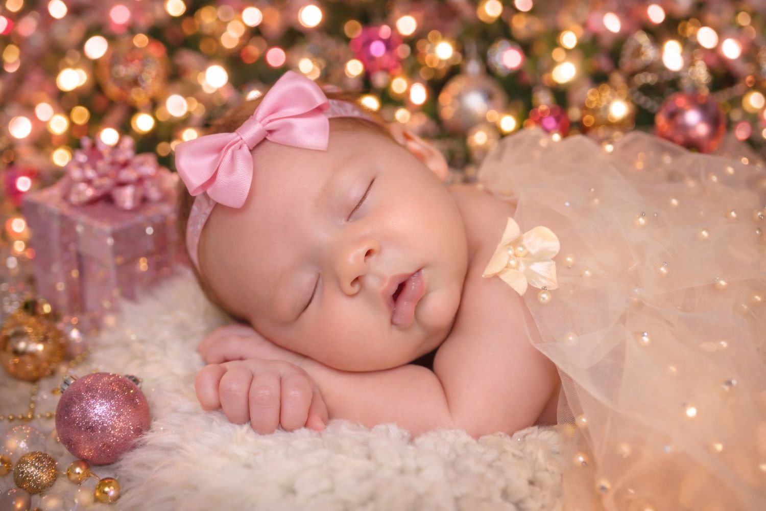 Sleeping baby in pink bow headband lying on soft white blanket with sparkling bokeh lights in background.