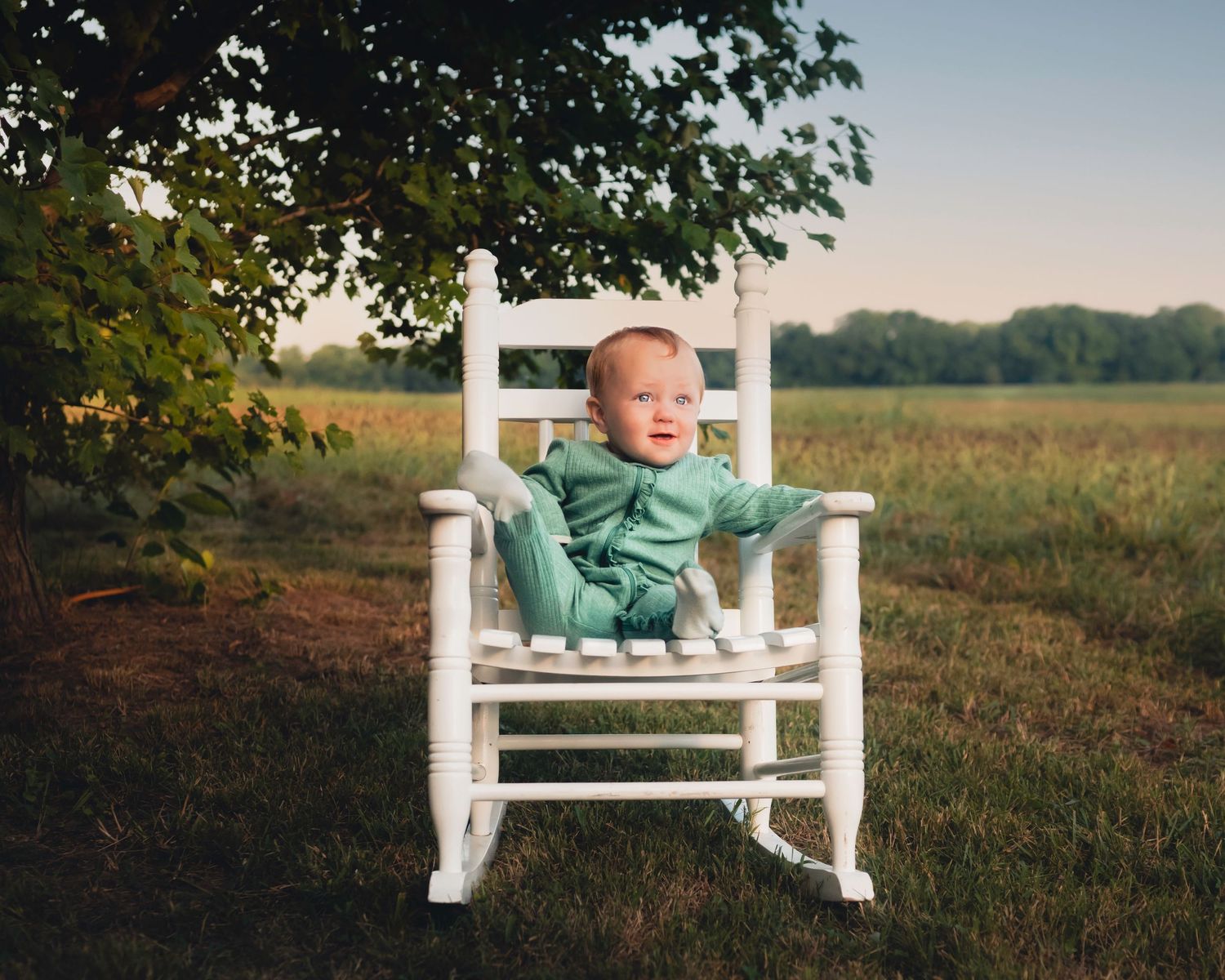 Baby in mint green outfit sits on white wooden bench in outdoor field setting.