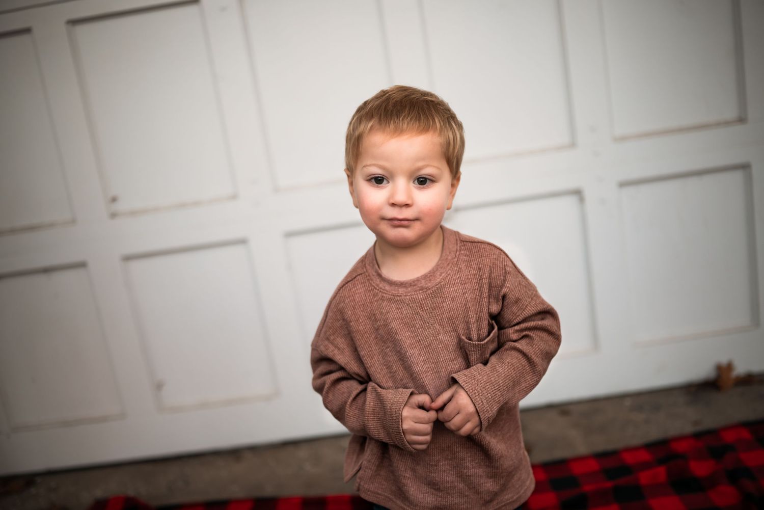 Small child wearing a brown sweater stands on a red and black plaid rug against a white paneled wall.
