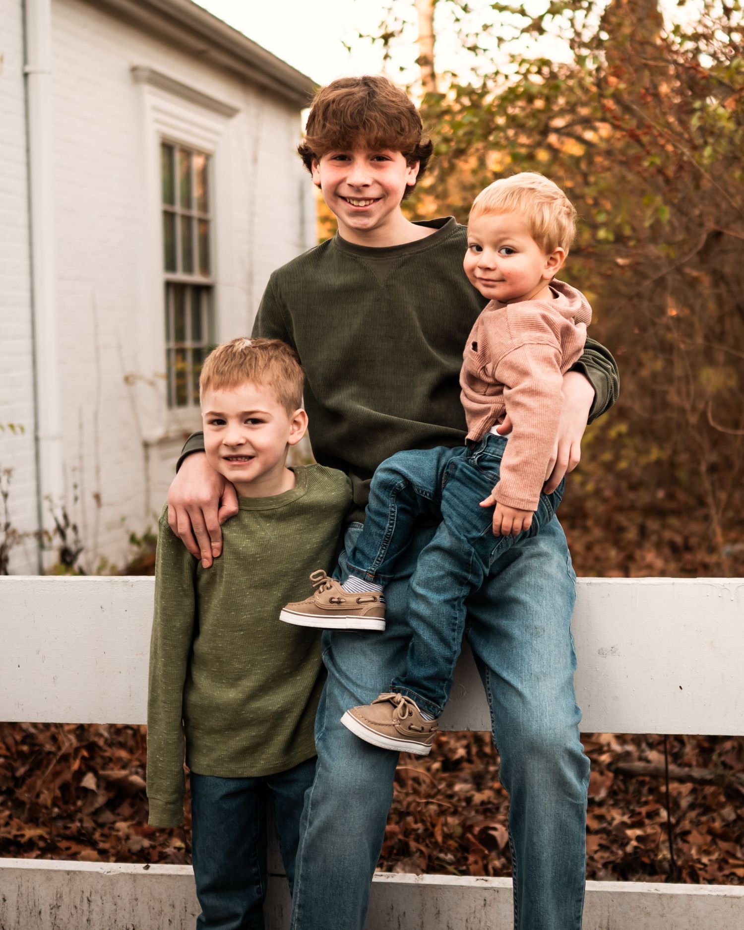 Three siblings pose together on a white fence wearing coordinating sweaters on an autumn day.