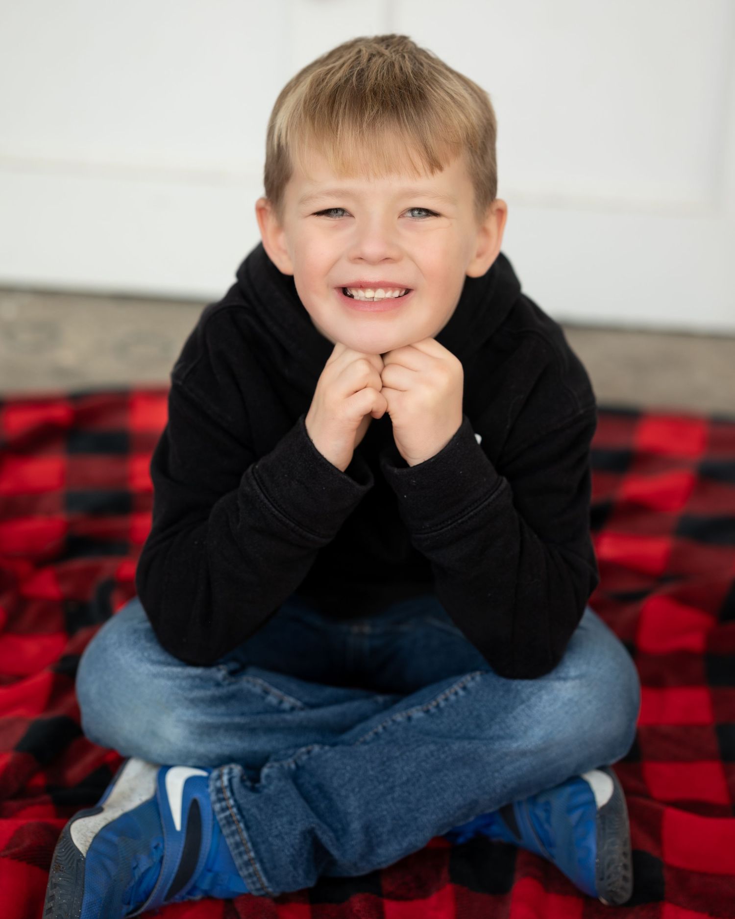 A child in a black sweater sits cross-legged on a red and black buffalo plaid blanket with a cheerful expression.