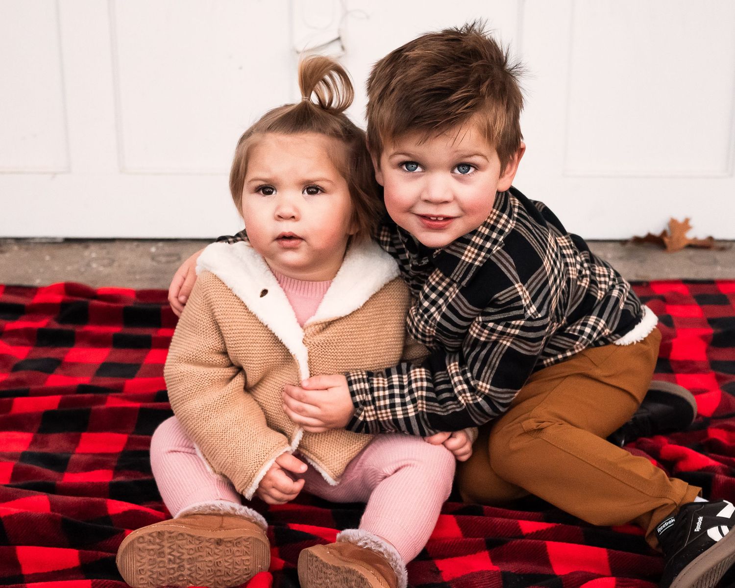 Two children in winter coats and scarves sit together on a red and black buffalo plaid blanket.