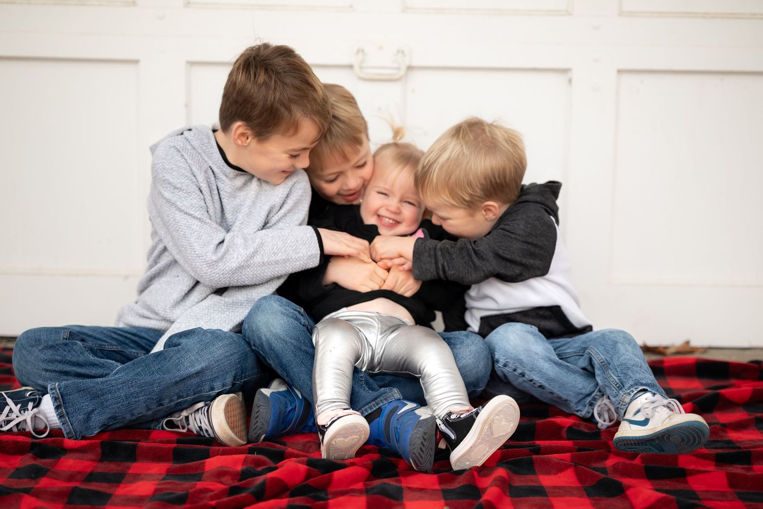 Children in matching gray sweaters and jeans sit together on a red and black buffalo plaid blanket.