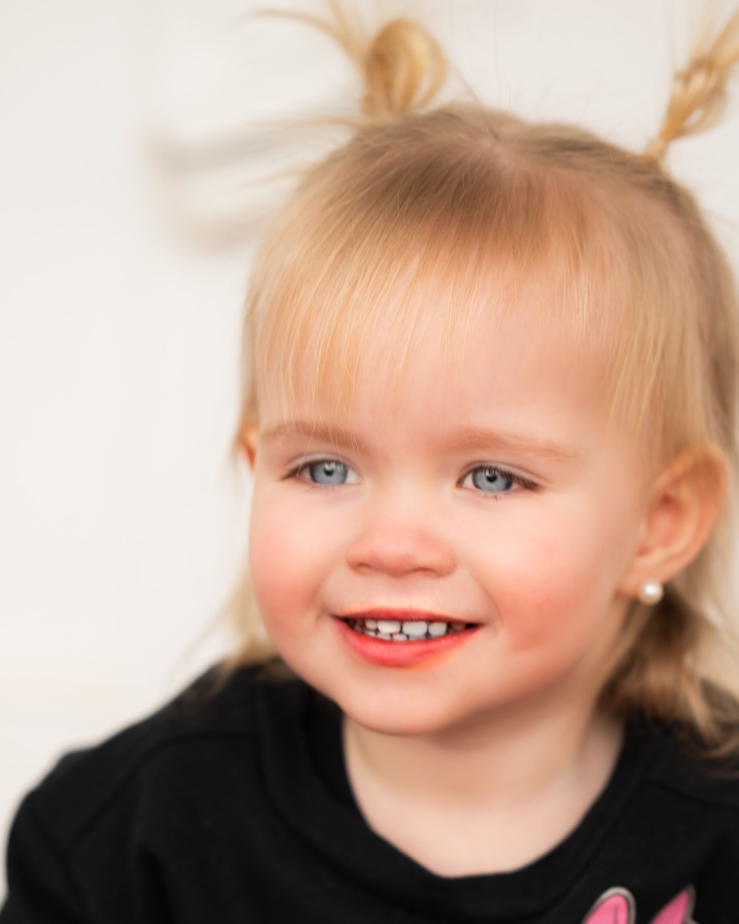 Young child in black shirt smiles at camera with bright blue eyes and blonde pigtails.