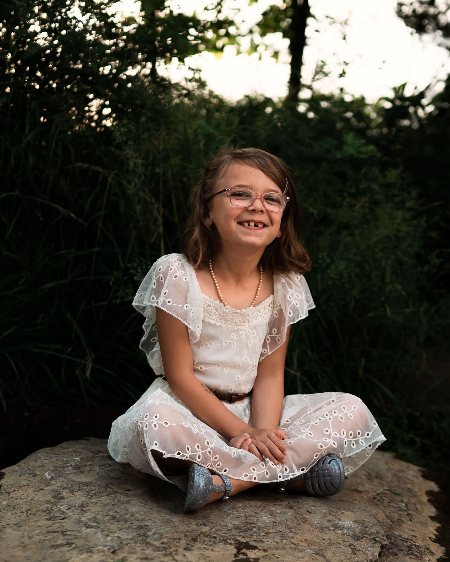 Young girl in white lace dress sitting on rock outdoors.