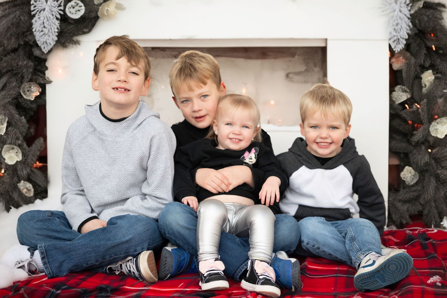 Four young siblings sit together on a red blanket in front of a Christmas tree wearing casual clothing.