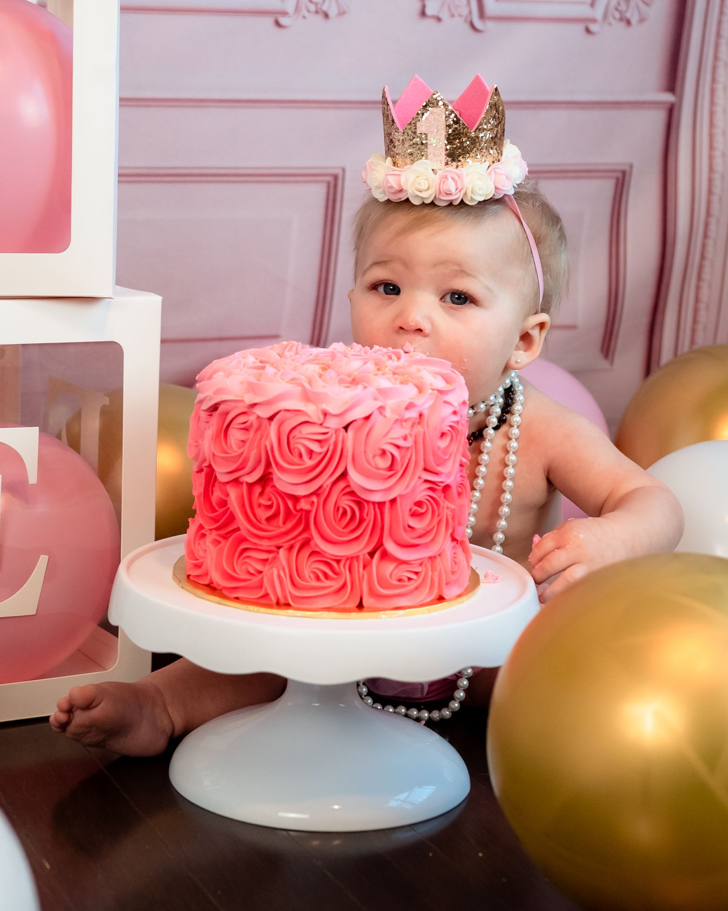 A pink rosette cake sits on a white pedestal stand surrounded by gold and pink balloons for a first birthday celebration.