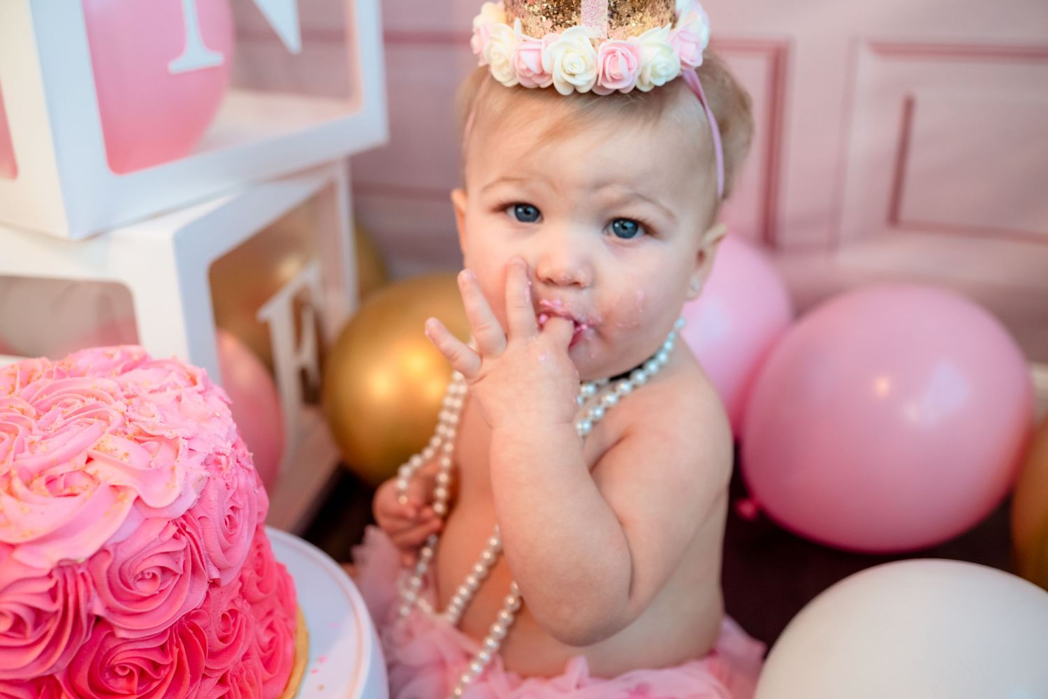 A pink rosette birthday cake with pearls and gold crown decorations sits among pink and gold balloons.