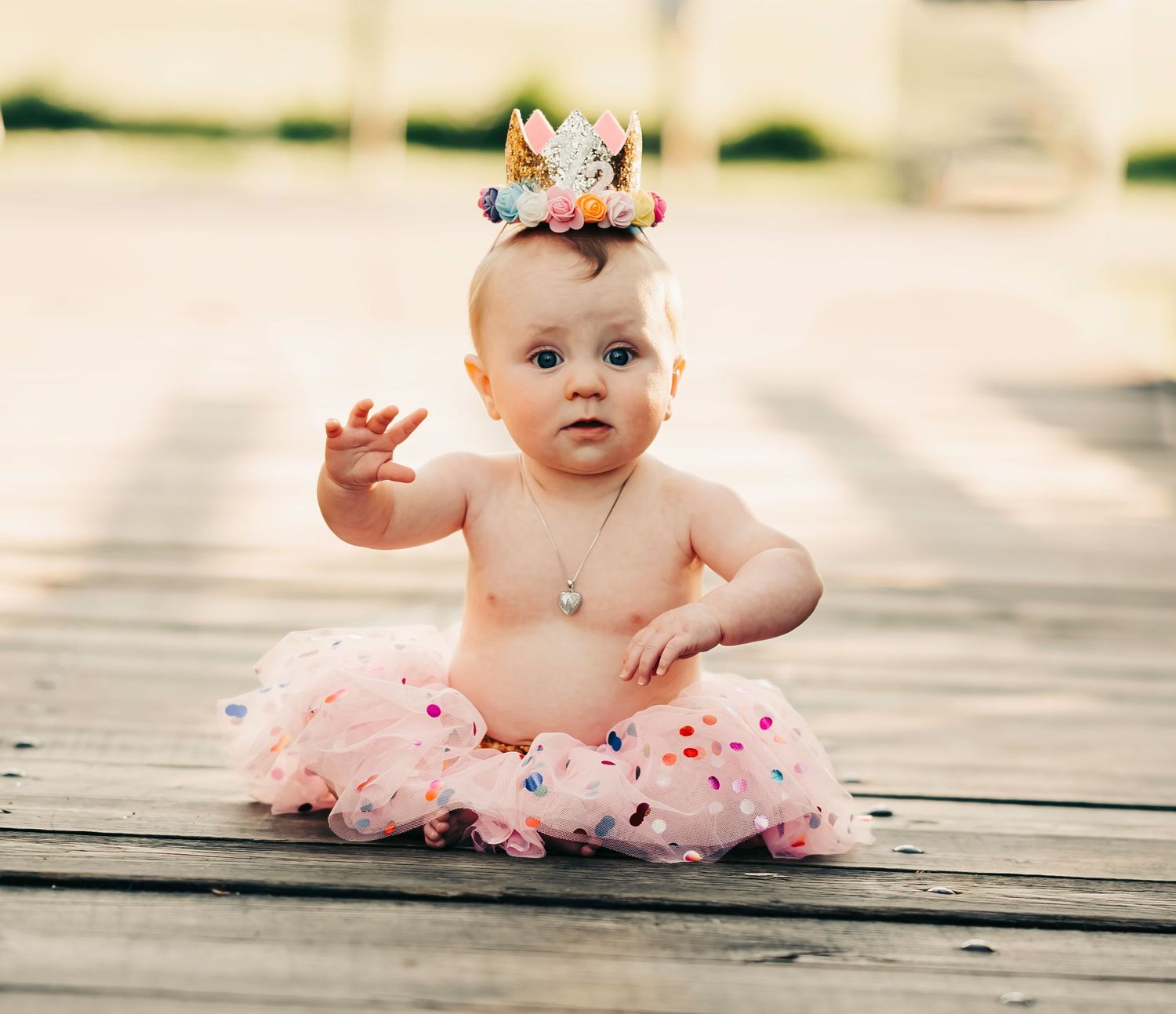 Baby wearing pink tutu sits on wooden deck.
