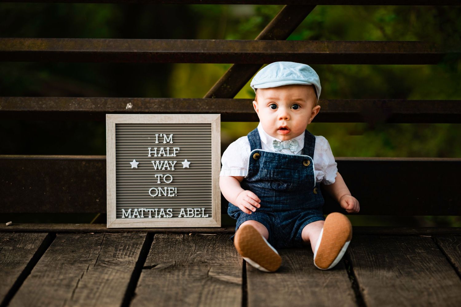 Baby in denim overalls sits with milestone board in outdoor setting.