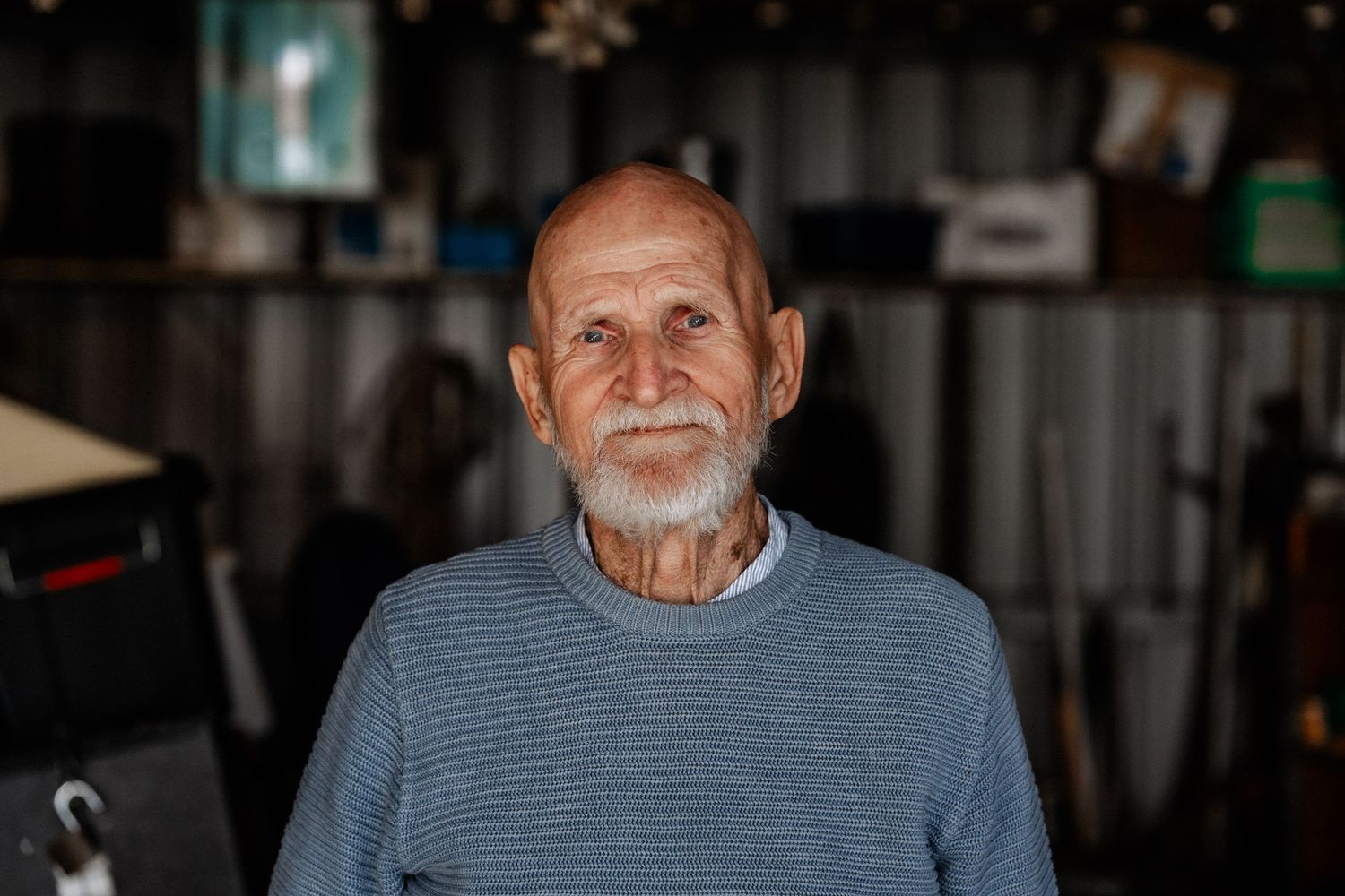 A portrait of an elderly man standing in a shed, looking directly at the camera.