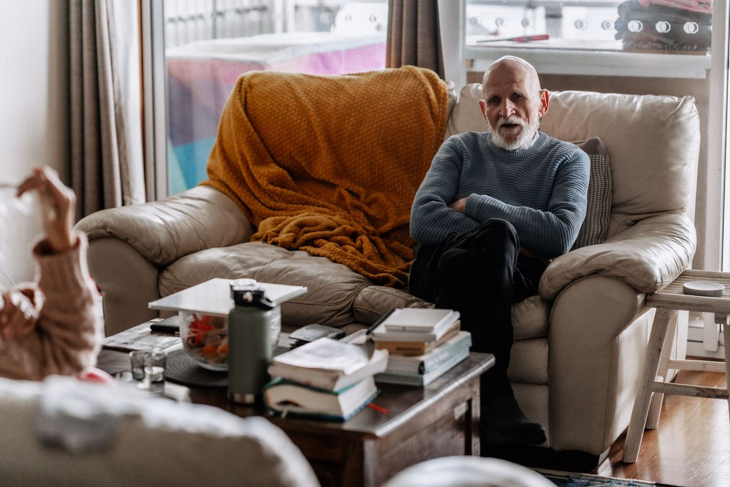An elderly man sits on a couch with his arms and legs crossed, looking at a coffee table.