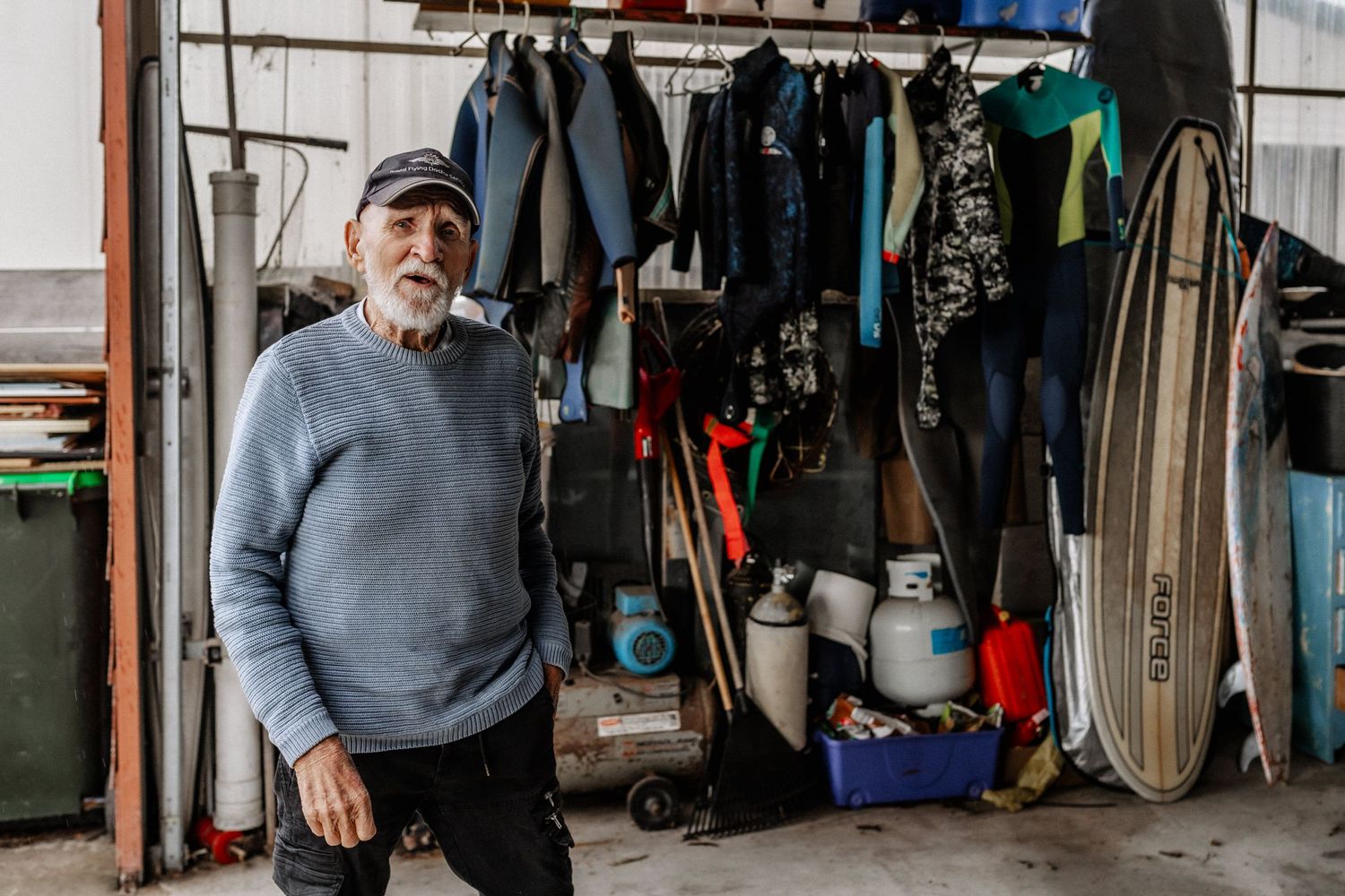 An elderly man stands with one hand in his pocket, in a garage, with wetsuits and surfboards behind him.