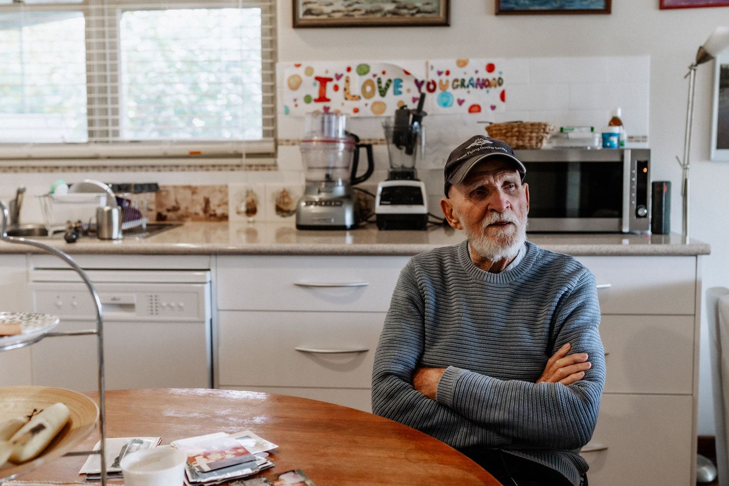An elderly man sits at a kitchen table with his arms crossed.