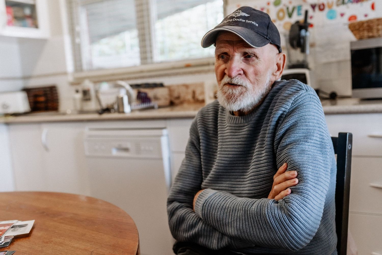 An elderly man tells a story while sitting at a kitchen dining table, with his arms crossed.