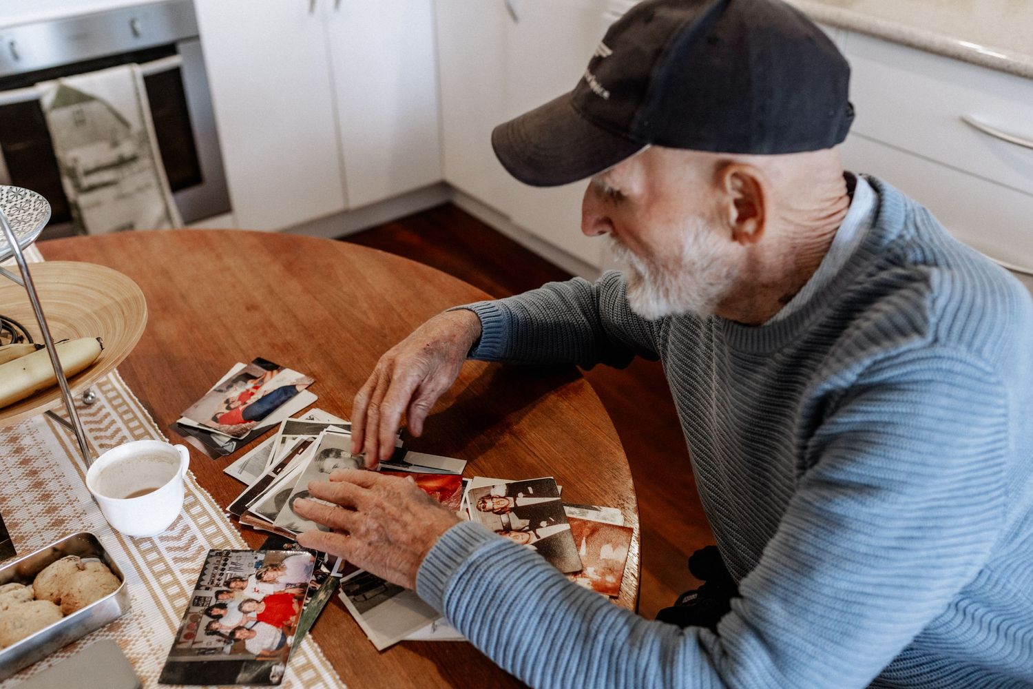 An elderly man sits at a kitchen table with old photos and a cup of tea.