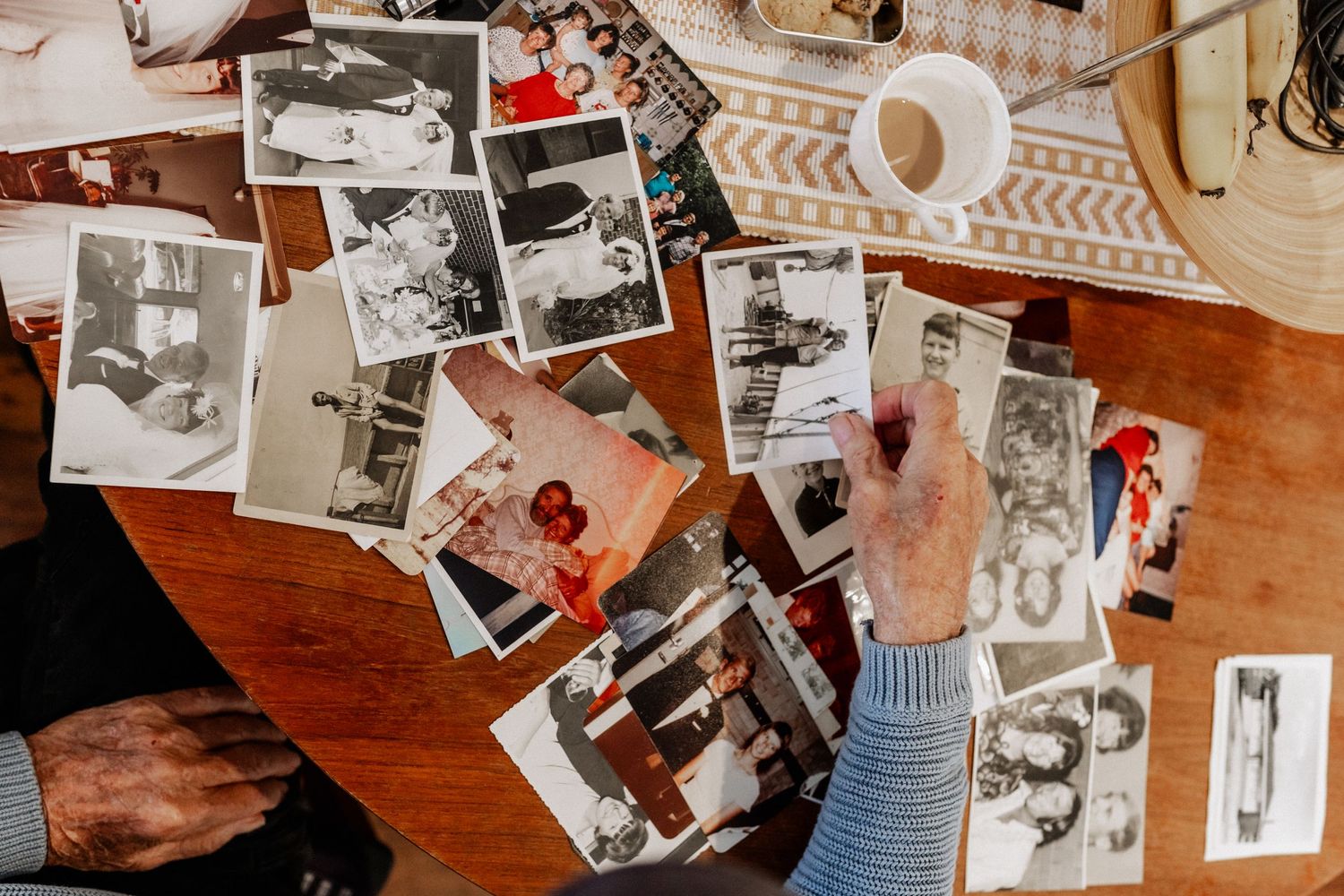 The hand of an elderly main reaching for an old photo, on a table full of old photos, momentos and a cup of tea.