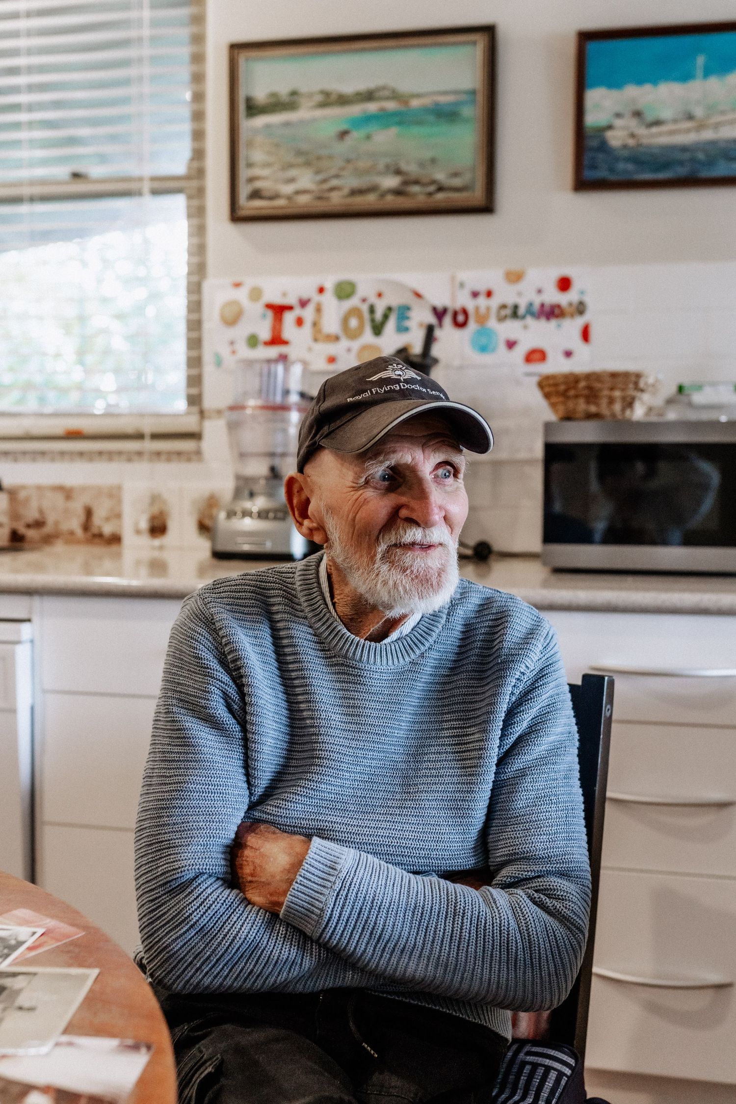 An elderly man sits at a dining chair in a kitchen with arms crossed, gazing into the distance.