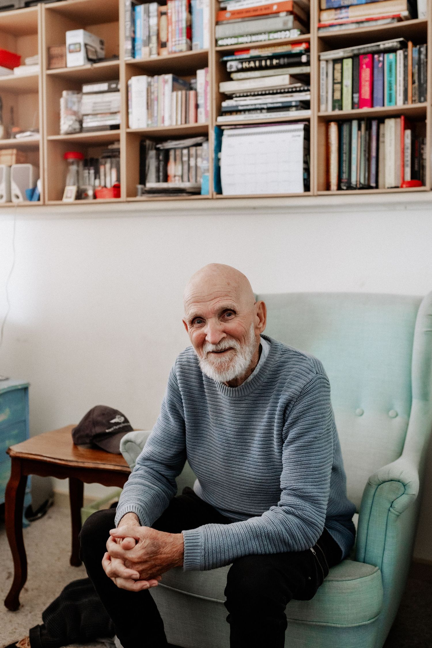 An elderly man sits in an arm chair, with his elbows on his knees. Bookcases are on the wall above him.