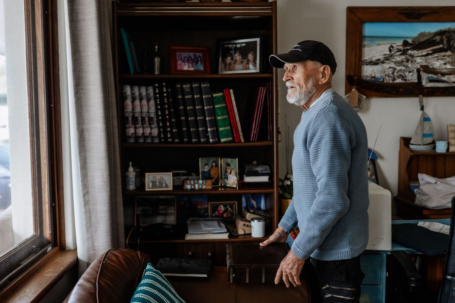 An elderly man gazes out of a window in a lounge room.