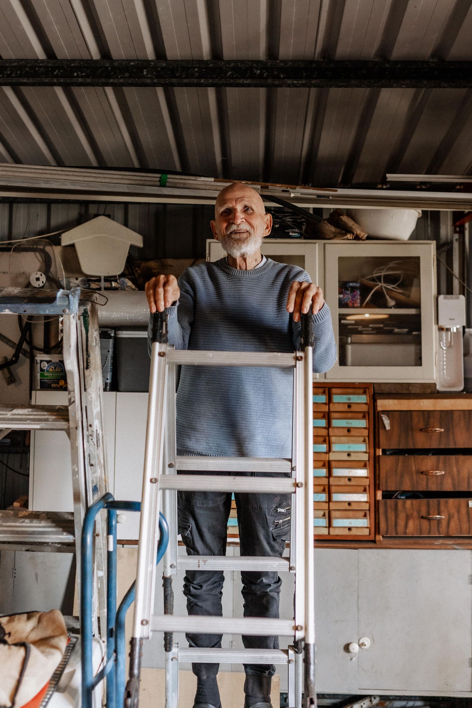 An elderly man stands on a ladder in a shed.