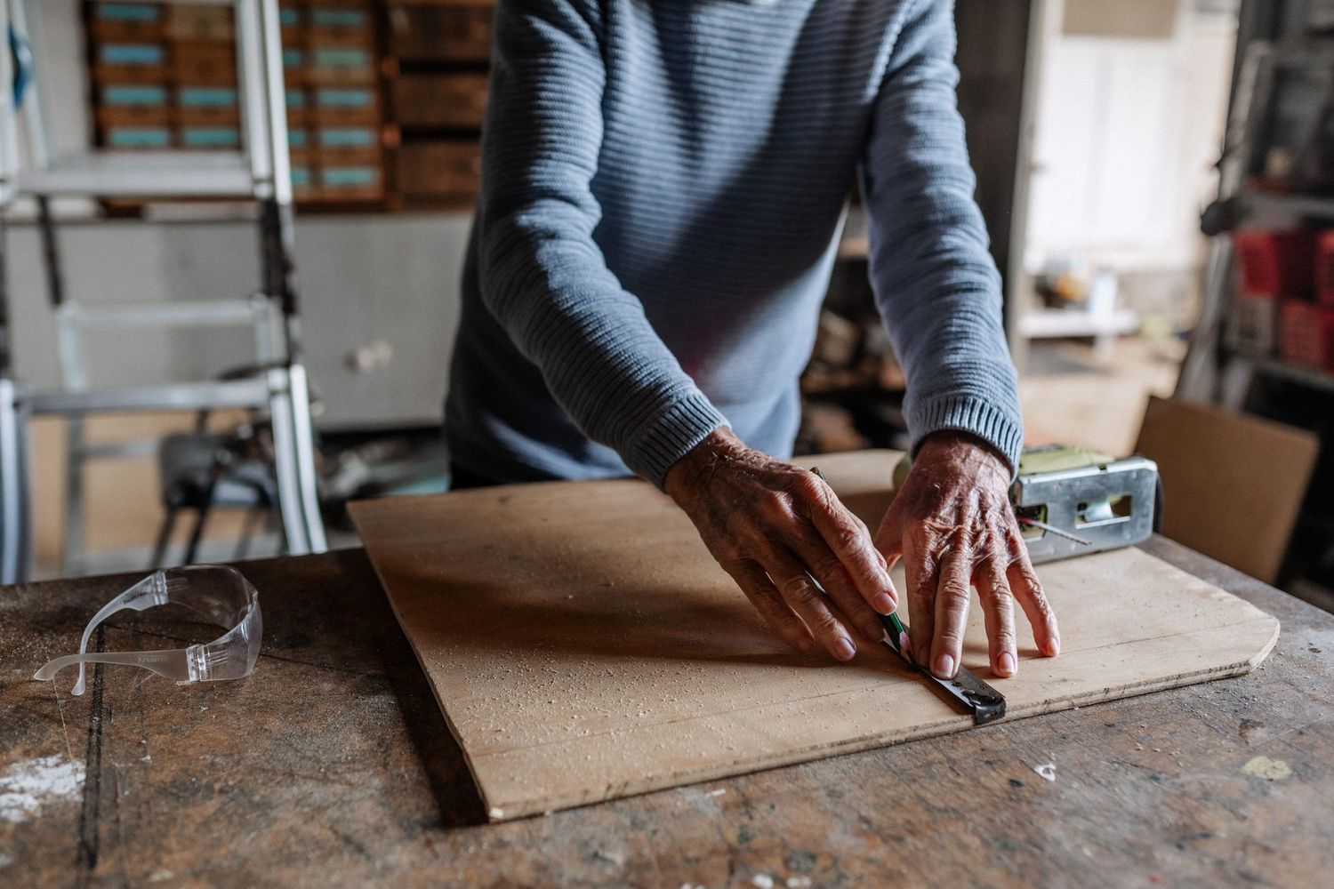 Hands of an elderly man in a shed, drawing a straight line on timber using a straight edge.