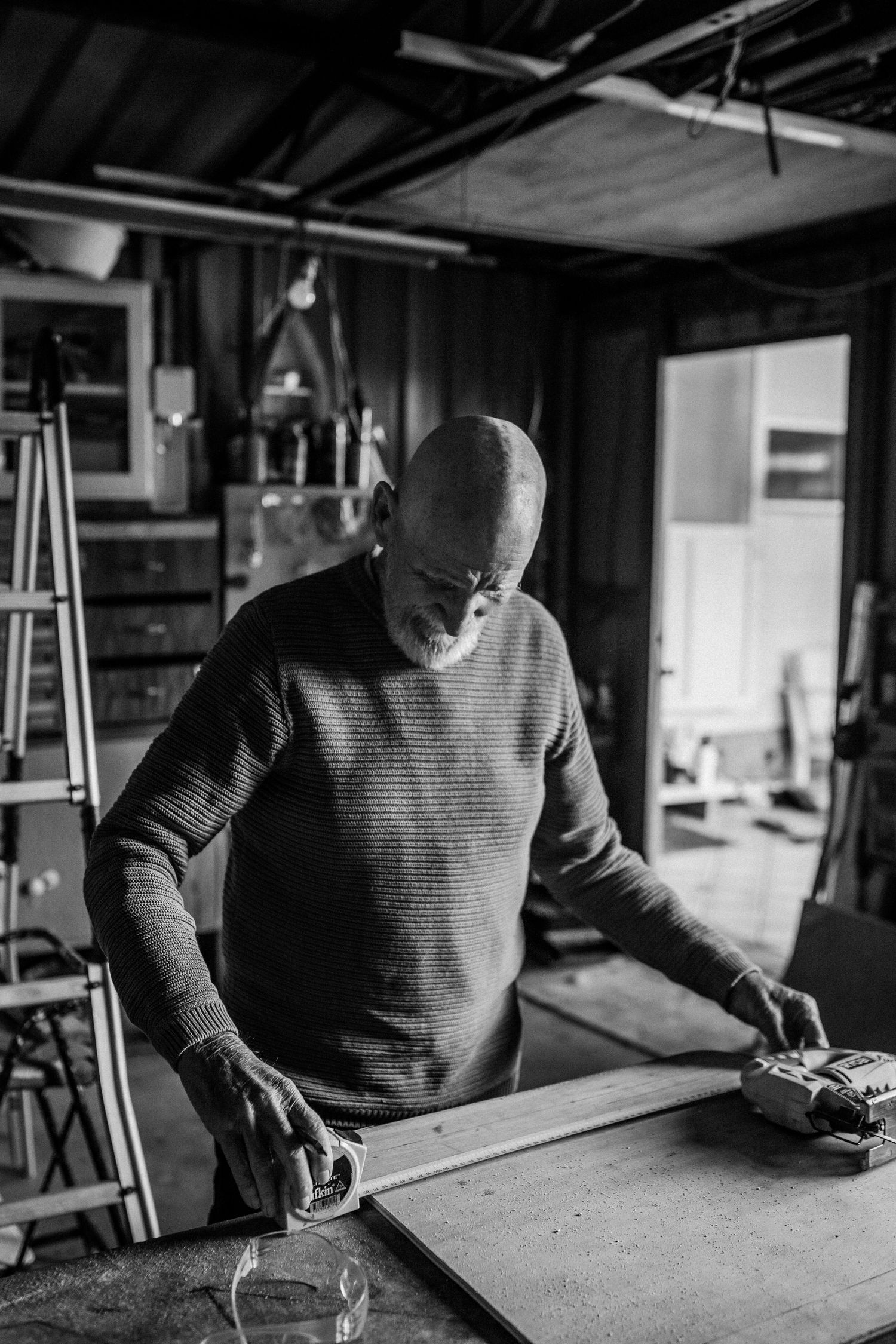 An elderly man in a shed, measuring a piece of wood with a tape measure.