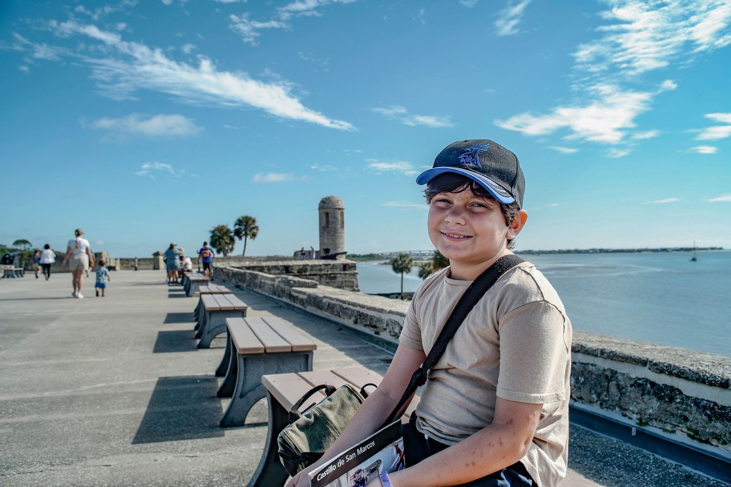 A sunny waterfront walkway with benches, palm trees and a historic tower visible in the distance.