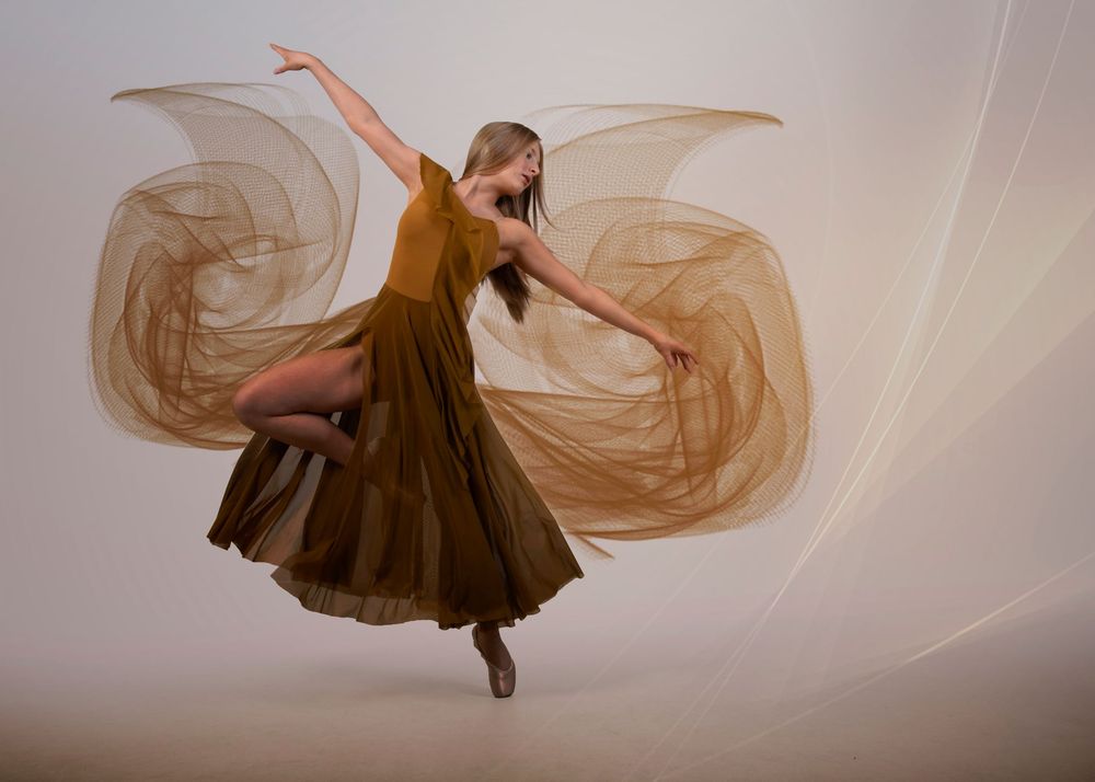 Graceful ballet dancer leaping in a flowing golden brown dress with fabric swirling dramatically against a neutral background.