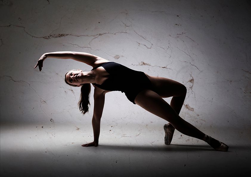 Modern dancer in black attire performs dramatic side pose in moody lighting.