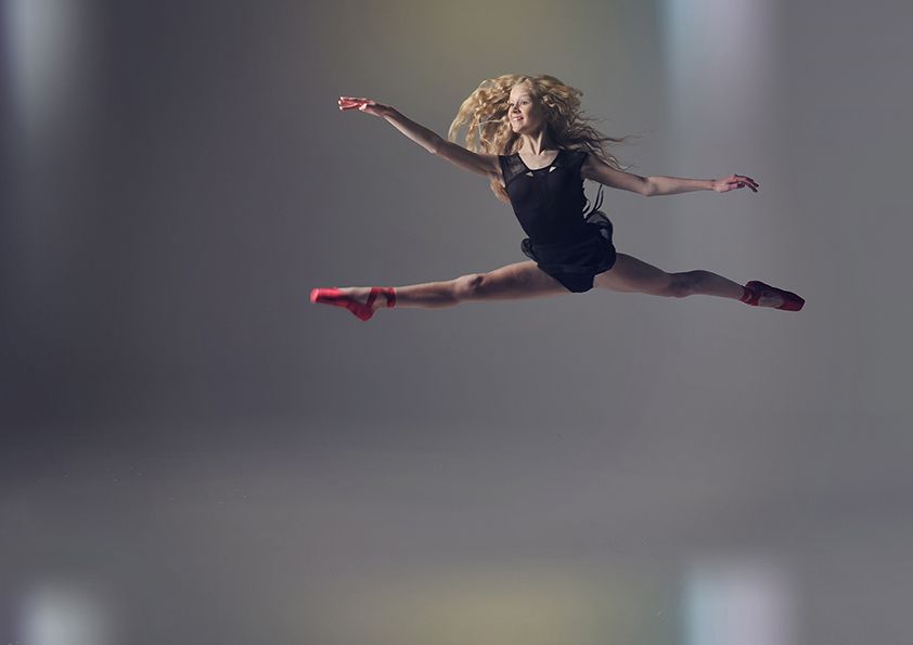 Dancer executes powerful split leap in black leotard and red pointe shoes.