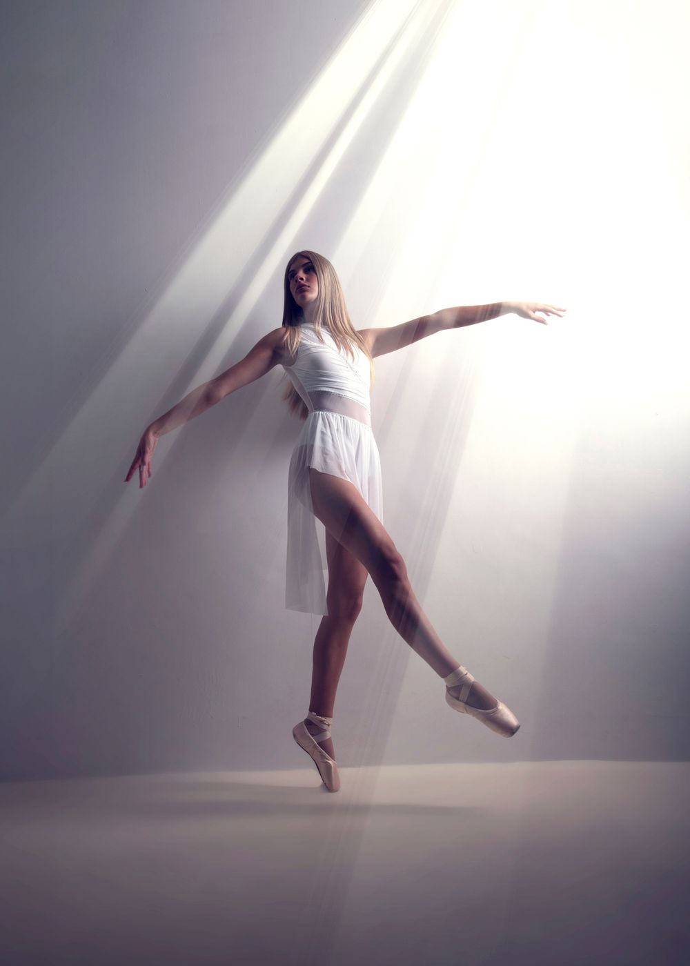 A graceful ballerina in white dress performs on pointe in a dramatic beam of light against a soft gray background.
