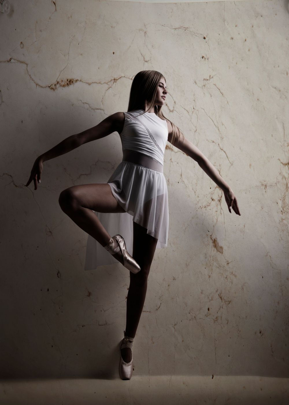 A graceful ballerina in a white tutu and pointe shoes poses elegantly against a textured marble wall.
