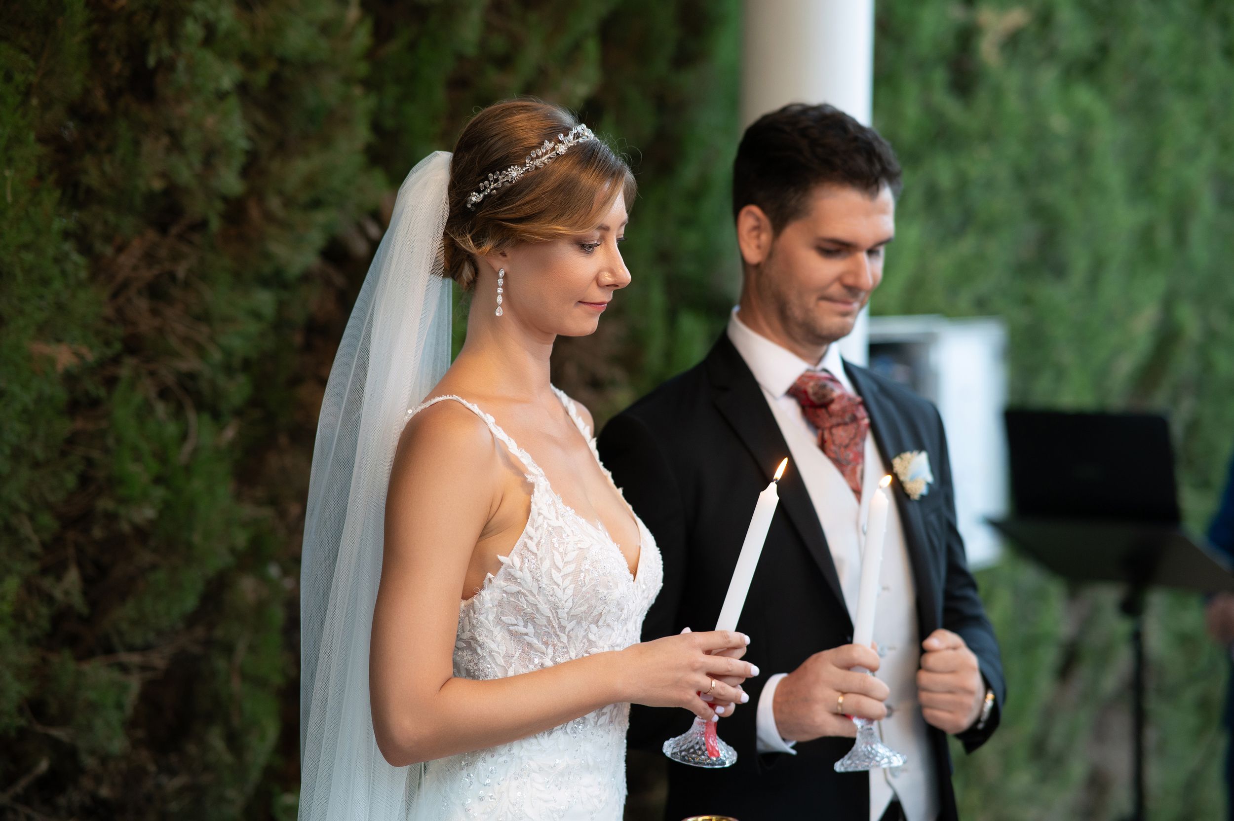A newlywed couple holds champagne glasses during an elegant outdoor wedding celebration with lush greenery in background.