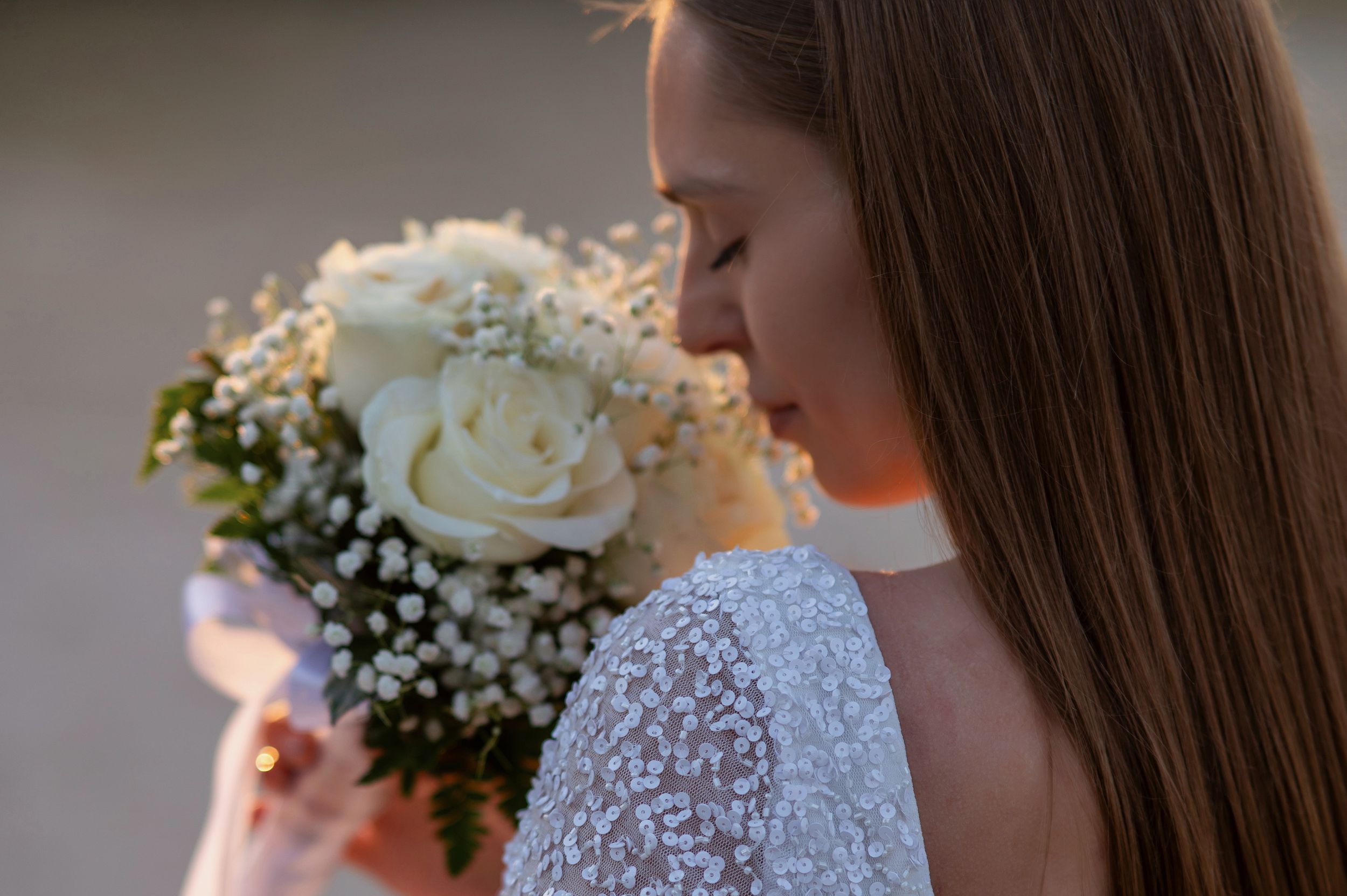 Side view of bride in white lace dress holding a bouquet of white roses and baby's breath flowers in sunlight.