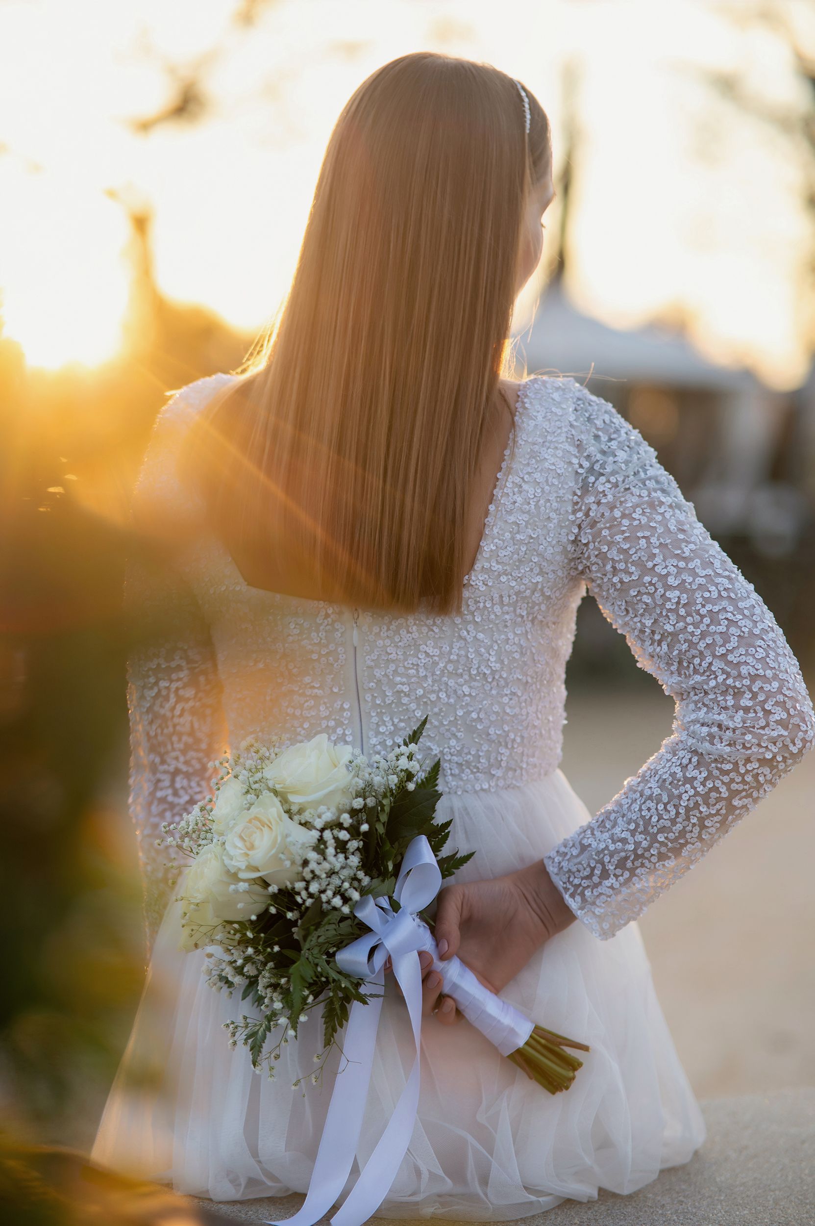 Back view of bride in lace sleeve wedding dress holding white bouquet during golden sunset hour.