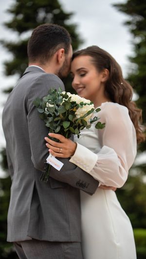 Romantic newlywed couple embracing outdoors with a white rose and eucalyptus bouquet against a garden backdrop.