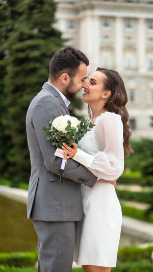 Intimate wedding moment as couple shares tender embrace while holding elegant white floral bouquet in garden setting.