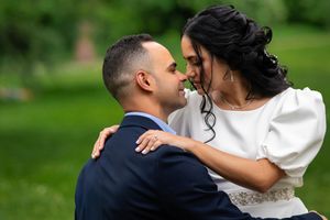 Couple in elegant wedding attire share an intimate moment outdoors with lush greenery in background.