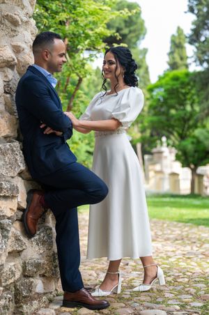 A couple in elegant attire share a romantic moment against a stone wall in a garden setting.