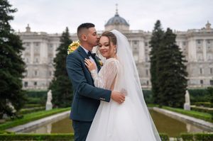 A wedding couple embraces in front of the Royal Palace of Madrid with its ornate architecture and manicured gardens.