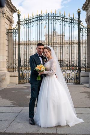 Wedding couple poses in front of ornate palace gates with yellow bouquet and elegant white dress and dark suit.