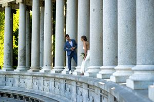 Two people stand between classical stone columns at an elegant outdoor venue with lush greenery in the background.