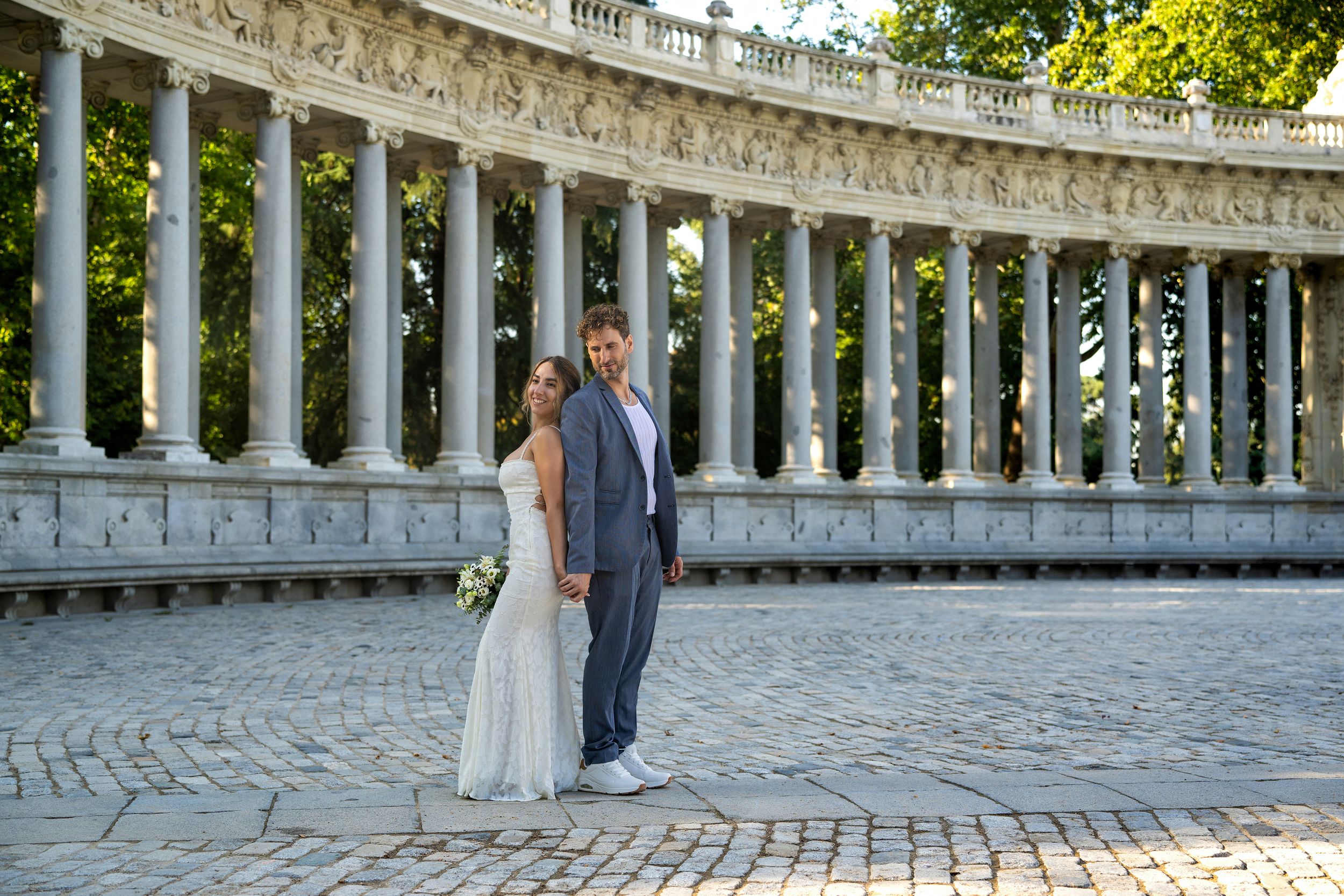 A couple in wedding attire stands in front of ornate white stone columns at Retiro Park in Madrid, Spain.