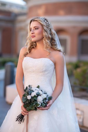A bride in a white strapless wedding gown holds a eucalyptus and white floral bouquet while wearing a crystal headpiece and veil.